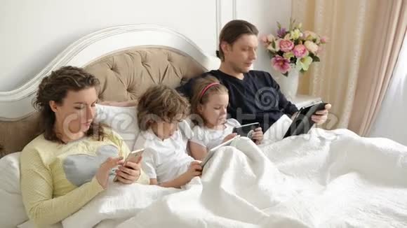 两个孩子和他们的父母一起度过周末躺在床上使用电子小工具现代视频的预览图