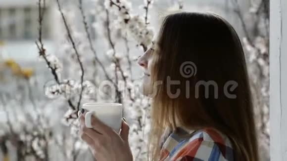女孩站在窗前的阳台上在盛开的杏树的背景下喝茶视频的预览图