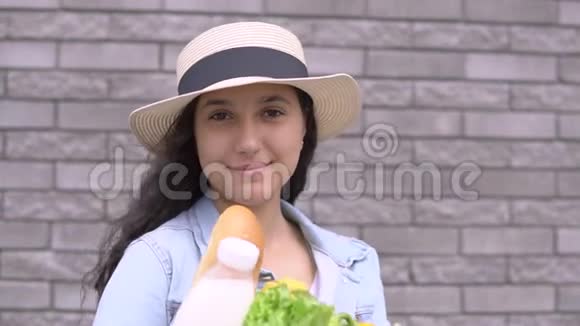 一个穿着牛仔夹克和帽子的年轻漂亮女孩抱着一袋蔬菜牛奶和面包心情很好视频的预览图