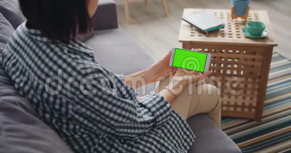 坐在家里沙发上看绿色智能手机模拟屏幕的女孩视频的预览图