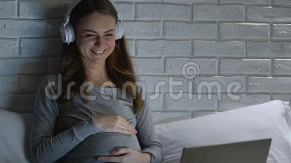 戴着耳机的怀孕女性笑着晚上在笔记本电脑上看喜剧表演视频的预览图