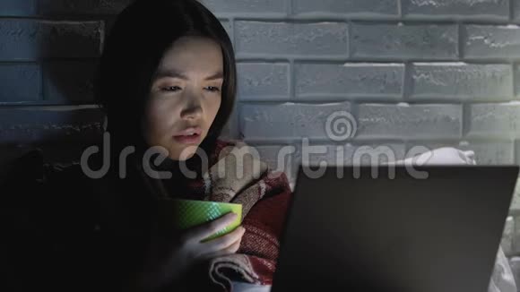 生病的女人被毯子覆盖晚上喝热茶在笔记本电脑上工作视频的预览图