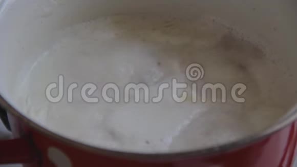 燕麦与牛奶厨师和煮沸在一个红色搪瓷锅在厨房炉子上泡沫聚集在表面煮熟搅拌孔视频的预览图