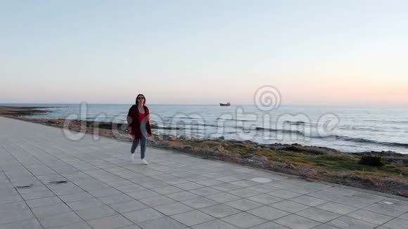 穿着太阳镜红色衬衫和牛仔裤的白种人黑发女孩在海滩上散步穿着红色泳衣蓝色牛仔裤的年轻迷人女子视频的预览图