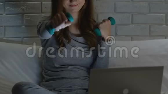 女人在床上用哑铃训练在笔记本电脑上看健身视频锻炼身体视频的预览图