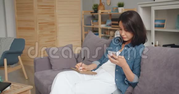 年轻女士在笔记本上做笔记在家里看智能手机屏幕视频的预览图