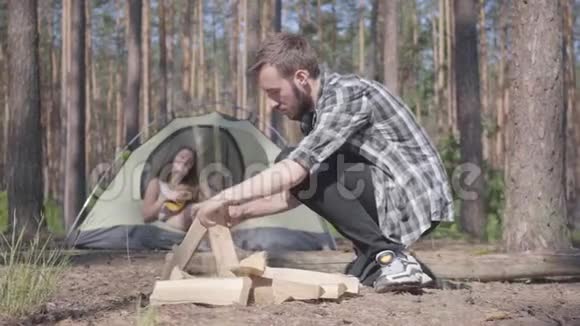 肖像帅哥穿着格子衬衫准备柴火在户外生火女孩坐在帐篷里玩视频的预览图