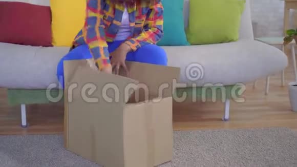 有着非洲发型的快乐而快乐的非裔美国妇女打开包裹坐在沙发上视频的预览图