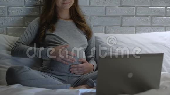 快乐的孕妇抚摸肚子晚上在笔记本电脑上看视频课视频的预览图