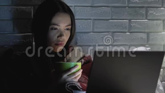 生病的女人拿着毯子在笔记本电脑前喝茶头疼视频的预览图