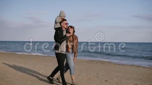 一个年轻时尚的戴墨镜的父亲把他的儿子扛在肩上一个年轻的家庭在海边散步享受关节视频的预览图