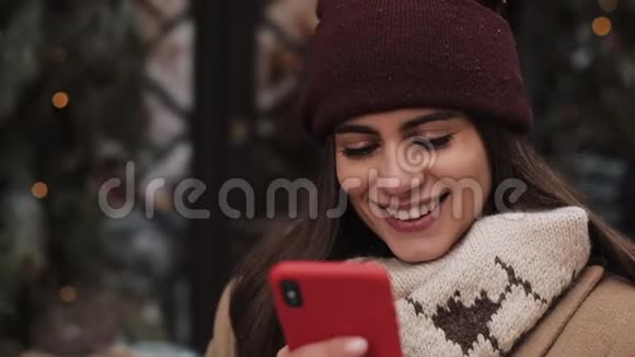 冬帽微笑女孩的特写和社交网络中的手套聊天使用她的智能手机看着视频的预览图