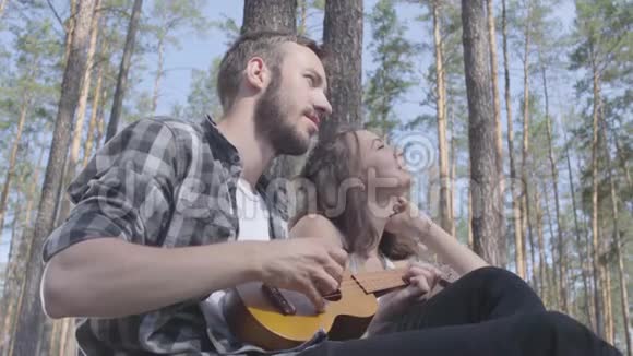 肖像吸引人的胡须年轻男子玩四弦琴而漂亮的年轻快乐的女人坐在附近可爱的一对视频的预览图
