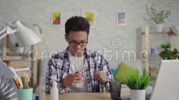 一个穿衬衫的亚裔年轻人坐在笔记本电脑旁用水冲洗药丸视频的预览图