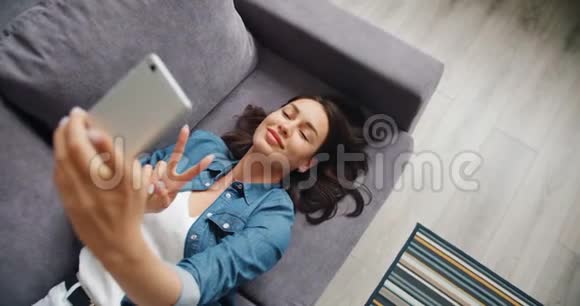 可爱的年轻女士在沙发上用智能手机自拍的俯视图视频的预览图
