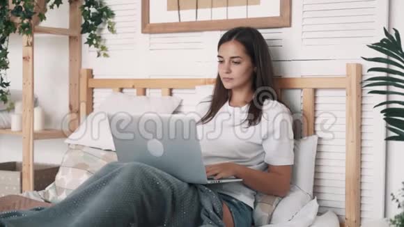 疲惫的女人坐在床上用笔记本电脑在家里工作打哈欠慢动作视频的预览图