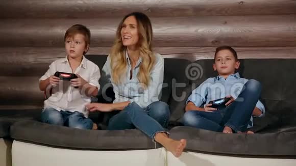 母亲和两个儿子坐在家里的沙发上用无线操纵杆玩电子游戏快乐的人们视频的预览图