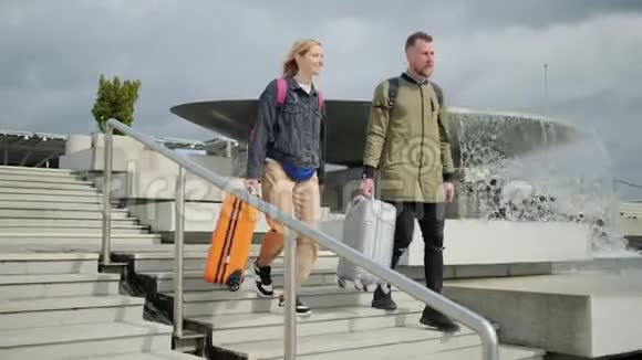 快乐的年轻人和女人带着手提箱从阳光明媚的街道上的楼梯上下来视频的预览图