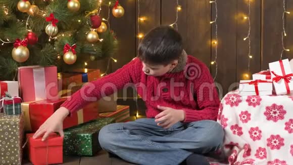 男孩坐在室内靠近装饰的圣诞树灯光和礼物圣诞快乐和节日快乐视频的预览图