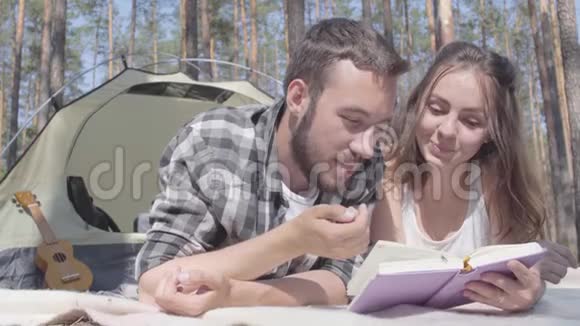 画像胡须男和可爱的年轻女子躺在森林里的帐篷里看书爱视频的预览图