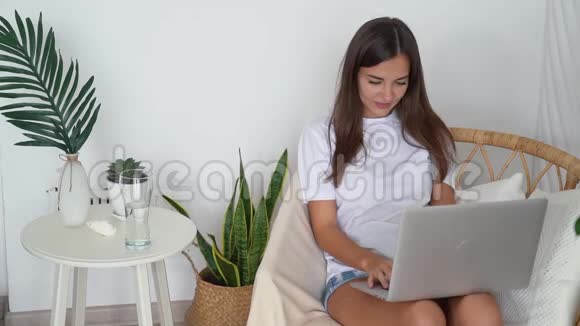 女孩坐在椅子上家里拿着笔记本电脑喝着玻璃里的清水慢动作视频的预览图