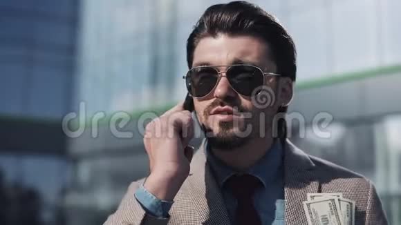 年轻英俊的商人穿着西装戴着太阳镜看上去很满意地在电话里拿着钱在附近说话视频的预览图