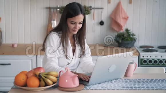 穿着浴袍的快乐黑发女人在厨房的笔记本电脑上工作视频的预览图