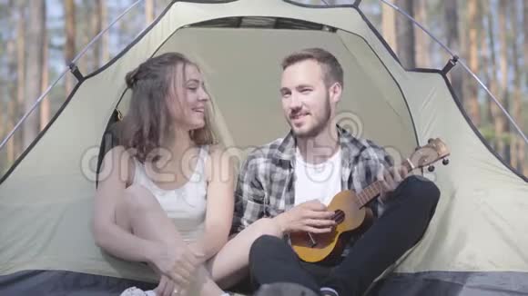 那个年轻人在帐篷里玩四弦琴而漂亮的年轻女人坐在森林里可爱的情侣玩得开心视频的预览图