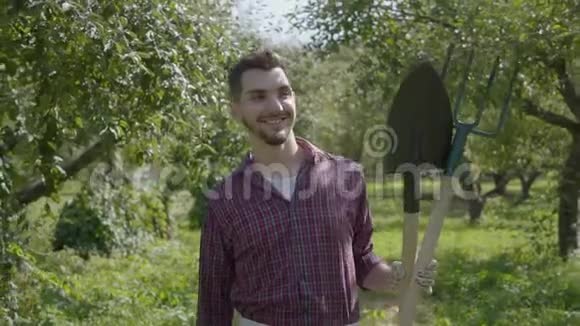 年轻的笑着留着胡子的农民手里拿着铲子和草叉穿过花园农村生活观念视频的预览图