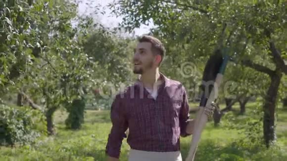 年轻的笑着留着胡子的白种人农民拿着铁锹和鹳雀在花园里查看果树视频的预览图
