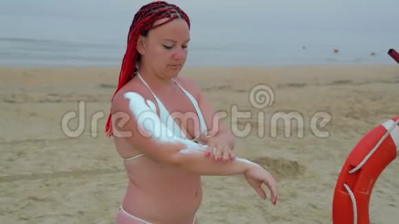 一位身穿白色泳衣的红发年轻女子正在海边给身体涂防晒霜视频的预览图