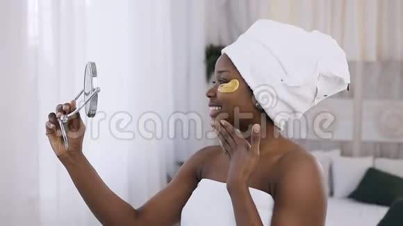 微笑着的非裔美国妇女眼睛下面贴着金色的补丁头上有白色的毛巾在镜子里看着在里面视频的预览图
