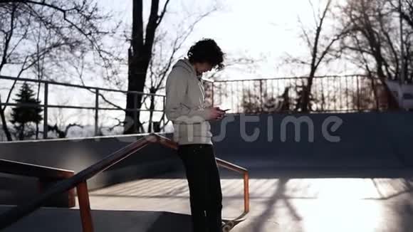 在阳光明媚的一天一个年轻的卷曲头男滑板运动员在滑板公园的手机里浏览精益视频的预览图