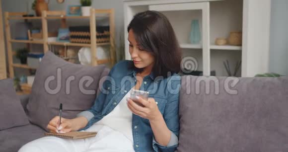 聪明的女人用智能手机在笔记本上写字微笑着坐在家里的沙发上视频的预览图
