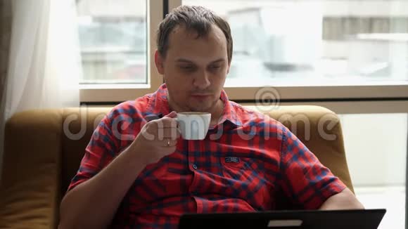 男人坐在现代客厅的沙发上用笔记本电脑工作在家喝咖啡视频的预览图