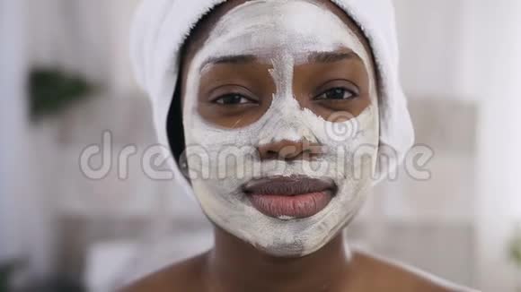 漂亮的美国黑人女孩头上戴着白色毛巾脸上戴着清洁面具看着镜头视频的预览图