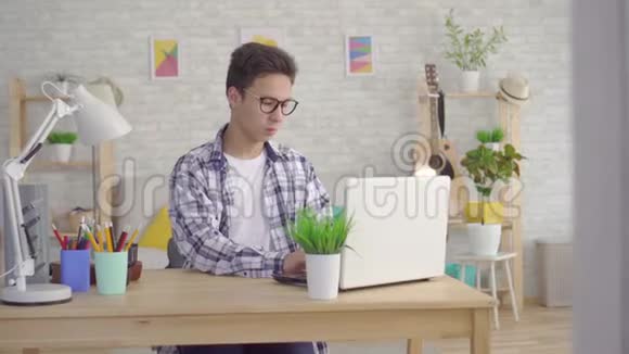 一个戴着时髦眼镜的亚洲年轻人赢得了胜利在家里客厅的笔记本电脑后面跳舞视频的预览图