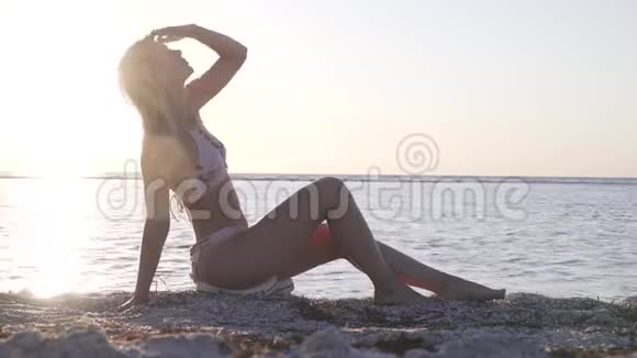 年轻的金发女郎穿着比基尼正坐在海滩上在日落时摆姿势视频的预览图