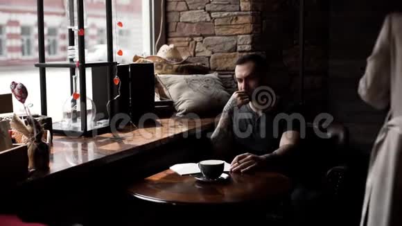 在咖啡馆里一个穿着纹身手臂坐在窗前的粗鲁男人在垫子上做笔记视频的预览图