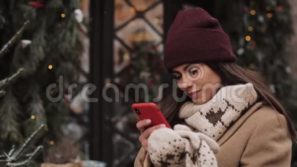 穿着冬帽和手套的漂亮白种人女孩使用她的智能手机看着屏幕微笑站在户外视频的预览图