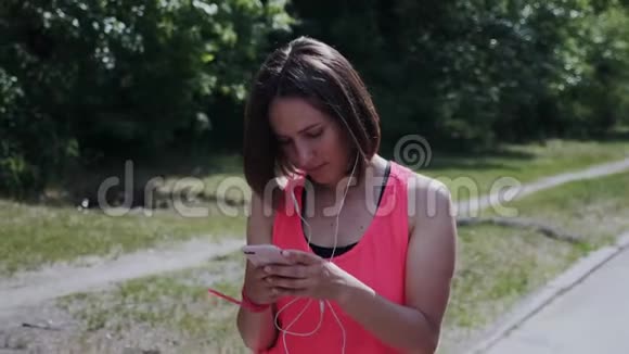 戴着耳机的可爱女孩穿过公园迷人的年轻女士戴着耳机在发短信看智能手机的女孩视频的预览图