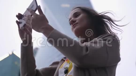 迷人的女人在建筑物和天空的背景下用手机自拍视频的预览图