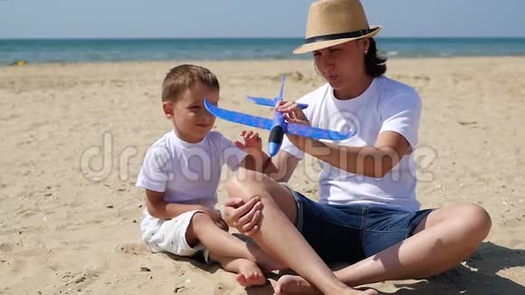 一个小可爱的男孩和他的妈妈在阳光明媚的夏日坐在沙滩上玩玩具飞机视频的预览图