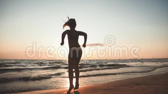 日落时在海里跳跃的少女剪影十几岁的女孩在海滩上的夕阳下跳舞暑假假期视频的预览图