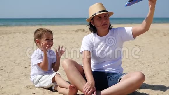 快乐的母亲和儿子在阳光明媚的夏日坐在沙滩上玩玩具飞机视频的预览图