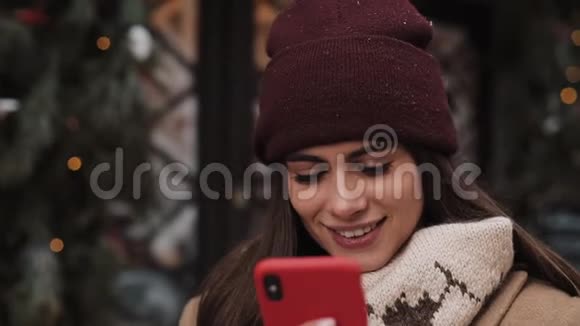 戴着冬帽和手套的微笑的漂亮女孩用她的智能手机看着屏幕聊天视频的预览图