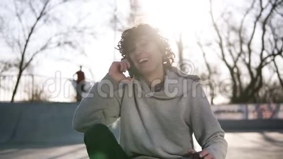 快乐微笑的年轻人正坐在溜冰公园的溜冰区用手机打电话给朋友一个男人视频的预览图