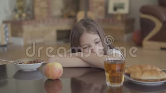 在上学前的清晨一个疲惫可爱的女孩正在muesli牛角面包和果汁附近吃早餐时睡着了视频的预览图