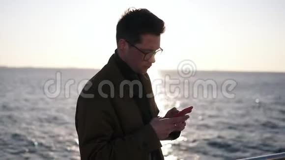 一位穿着棕色外套戴着时髦眼镜穿着智能手机发短信或在网上冲浪的白种人的肖像视频的预览图