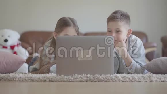 肖像可爱的双胞胎男孩和女孩躺在地毯上看着电脑上有趣的东西笑着视频的预览图